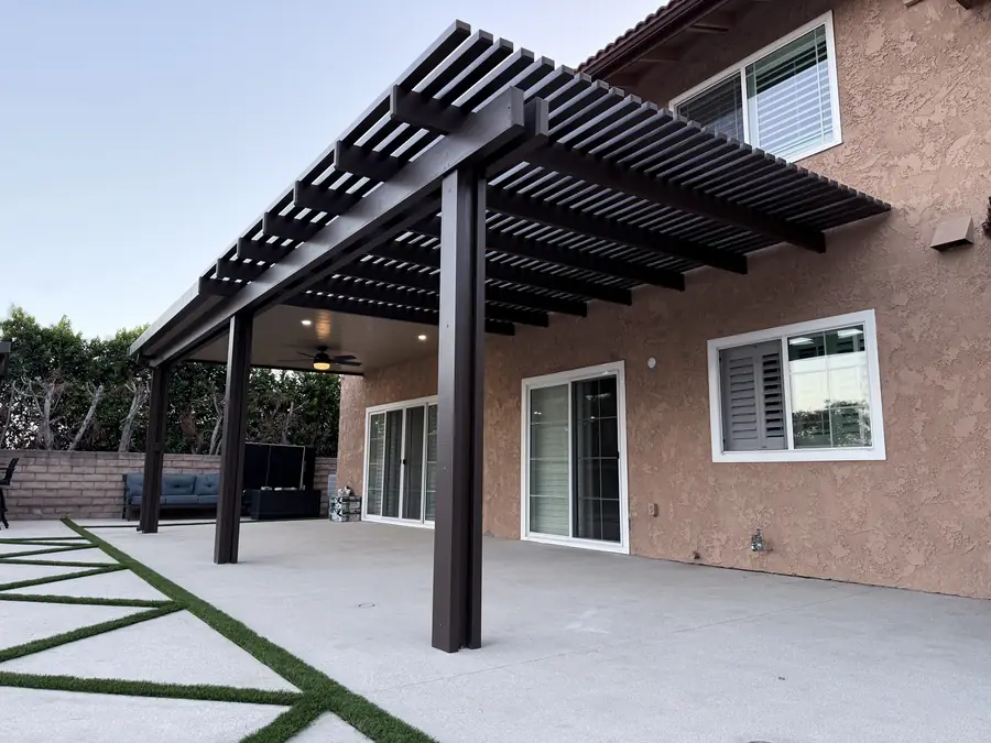 Dark brown aluminum lattice patio cover attached to a two-story stucco home over a concrete patio with turf strips
